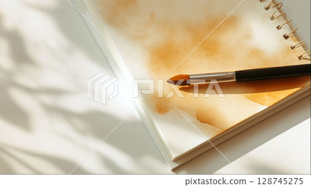 Watercolor brush resting on a spiral sketchbook with orange paint on textured paper in warm natural light 128745275