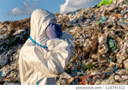Worker in protective gear surveys a large accumulation of plastic waste at a landfill under a clear blue sky during daylight 128745312