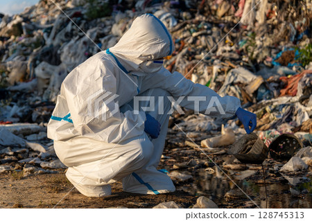 A dedicated environmental worker kneels beside a polluted area in a landfill, carefully collecting samples of contaminated water 128745313