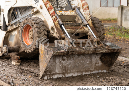 A skid steer loader with a dirt-filled bucket actively works on a muddy construction site, preparing for new building projects A skid steer loader with a dirt-filled bucket actively works on a muddy construction site, preparing for new building projects 128745320