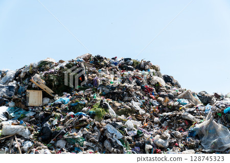 Piles of plastic and various waste materials fill the landfill under a clear blue sky. Littered debris and garbage are visible throughout 128745323