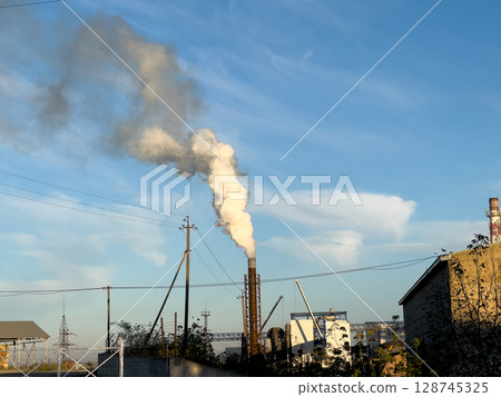Factory smokestack emits thick clouds of smoke into the bright afternoon sky, highlighting industrial activity and environmental concerns 128745325