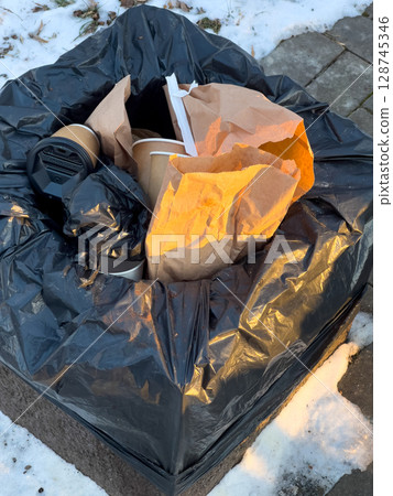 A large black garbage bag filled with paper and plastic waste sits on a snowy pavement, highlighting littering in a neighborhood A large black garbage bag filled with paper and plastic waste sits on a snowy pavement, highlighting littering in a neighborhood 128745346