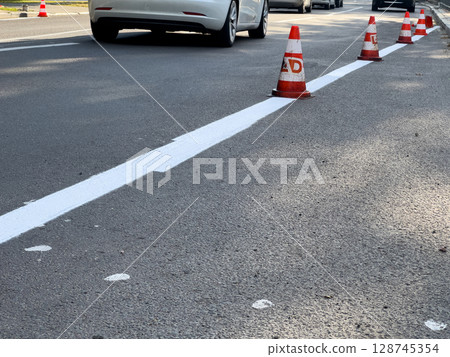 Traffic cones mark freshly painted white line on asphalt, indicating ongoing road work in an urban setting during the day Traffic cones mark freshly painted white line on asphalt, indicating ongoing road work in an urban setting during the day 128745354