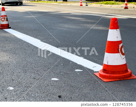 Traffic cones are positioned along a freshly painted lane marking in an urban area during road maintenance work, ensuring safety for drivers Traffic cones are positioned along a freshly painted lane marking in an urban area during road maintenance work, ensuring safety for drivers 128745356