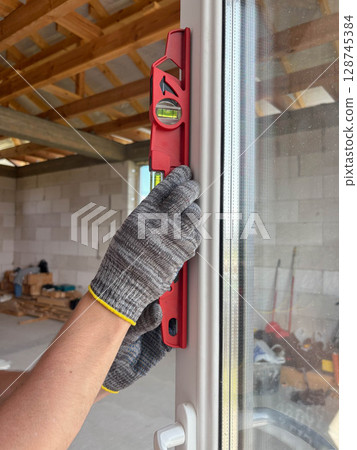 A worker wearing gloves and using a level checks the evenness of a new window at a construction site. A worker wearing gloves and using a level checks the evenness of a new window at a construction site. 128745384
