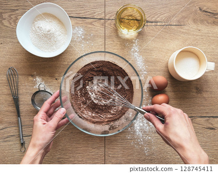 Female hands preparing chocolate sponge cake. Glass bowl with cocoa and flour on wooden table. 128745411