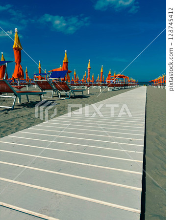 White wooden walkway along beach umbrellas and sun loungers on a sandy beach on a sunny day. 128745412