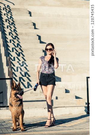A young girl wearing shorts and stylish glasses stands near a large staircase, holding a shepherd dog on a leash 128745435