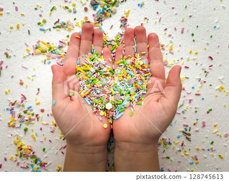 Sugar fine confectionery sprinkles form a heart shape in the palms of a child. Conceptual photo on a white background. 128745613