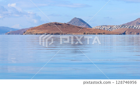 A serene seascape with rippling blue water in the foreground, leading to a rocky island and a distant coastal town nestled amidst arid mountains under a clear sky. High quality photo A serene seascape with rippling blue water in the foreground, leading to a rocky island and a distant coastal town nestled amidst arid mountains under a clear sky. High quality photo 128746956