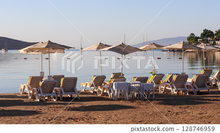 Beach chairs and umbrellas line a sandy shore, facing a calm sea with boats and distant hills under a clear sky. High quality photo 128746959