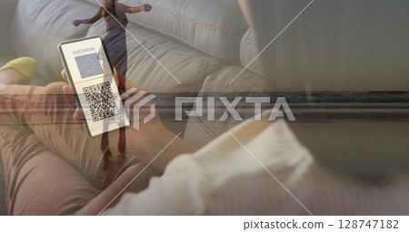 Image of woman with smartphone with covid passport over woman walking on beach 128747182