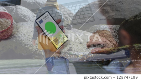 Image of woman with covid passport on smartphone over woman walking on beach 128747199