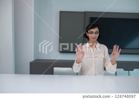 Asian woman wearing blouse sitting behind table in boardroom with flat-screen monitor and cabinets 128748097