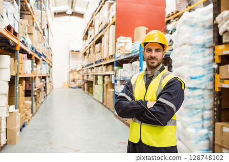 Male warehouse worker wearing hard hat in warehouse aisle next to stacked boxes, copy space Male warehouse worker wearing hard hat in warehouse aisle next to stacked boxes, copy space 128748102