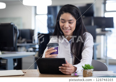 Hispanic female wearing headset while holding coffee cup and using tablet at desk beside succulent 128748103