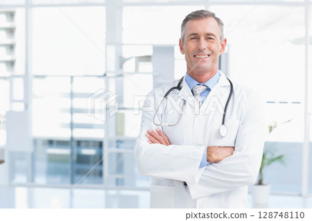 Mature male doctor smiling with stethoscope, crossed arms in clinic beside potted plant, copy space 128748110