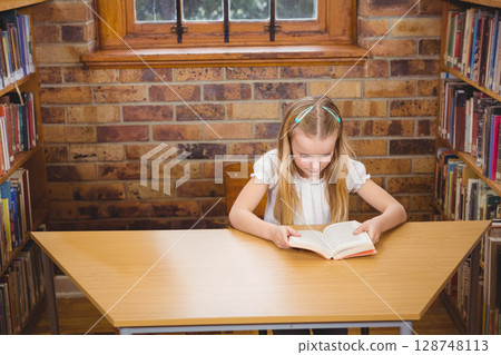 Girl reading paperback book at wooden desk in library with exposed brick wall and bookshelves Girl reading paperback book at wooden desk in library with exposed brick wall and bookshelves 128748113