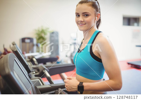 Fair-skinned woman standing beside treadmill in gym wearing earbuds and watch by plant, copy space Fair-skinned woman standing beside treadmill in gym wearing earbuds and watch by plant, copy space 128748117
