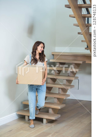 Mid adult woman carrying cardboard moving box while climbing wooden staircase in home stairwell 128748133