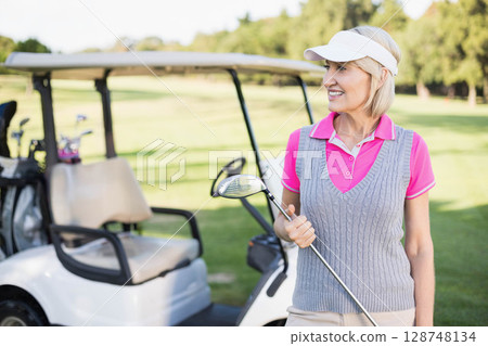 Senior woman wearing pink polo standing on fairway next to golf cart holding driver, copy space 128748134