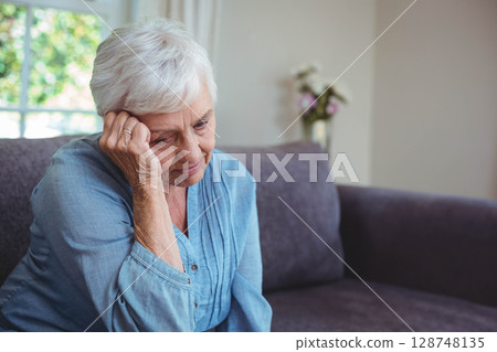 Senior woman in blue blouse sitting on grey sofa in living room gazing through sheer curtains 128748135