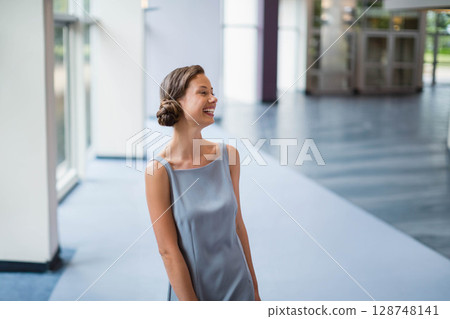 Mid-20s woman standing in modern lobby wearing satin dress by columns, glass doors, and windows Mid-20s woman standing in modern lobby wearing satin dress by columns, glass doors, and windows 128748141