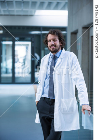 Male doctor standing wearing white lab coat, dress shirt and tie near corridor railing, glass doors 128748142