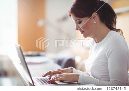Woman typing on laptop with notebook on home office desk near window with desk lamp Woman typing on laptop with notebook on home office desk near window with desk lamp 128748145