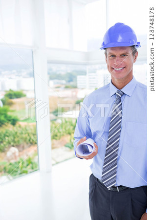 Man standing in office wearing blue hard hat and holding rolled plans by window, copy space 128748178