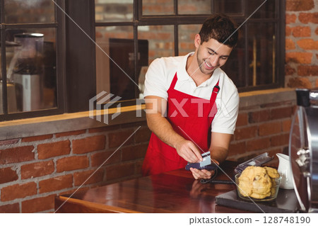 Male barista wearing apron handling payment at cafe counter by payment terminal and credit card 128748190