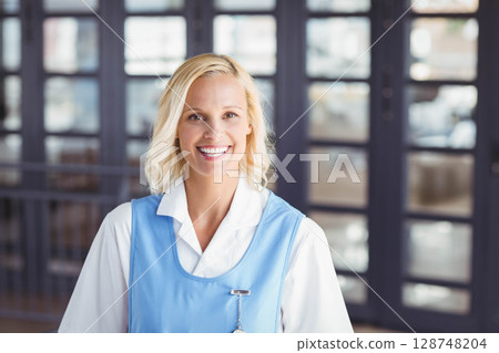Middle-aged female hotel staff member standing in blue smock at lobby doors with name badge 128748204