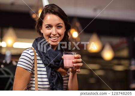 Woman standing in cafe under lamps holding cup of pink drink with straw and tan-strap bag 128748205
