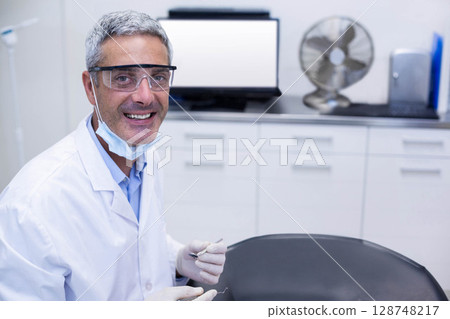 Male dentist wearing lab coat and goggles holding dental mirror and probe in operatory, copy space Male dentist wearing lab coat and goggles holding dental mirror and probe in operatory, copy space 128748217