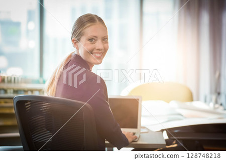 Businesswoman smiling and working at office desk by large windows with laptop and blueprints 128748218
