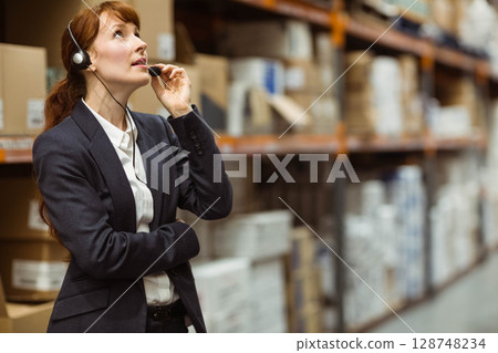 Female supervisor checking boxes on pallets while adjusting headset mic in warehouse, copy space Female supervisor checking boxes on pallets while adjusting headset mic in warehouse, copy space 128748234