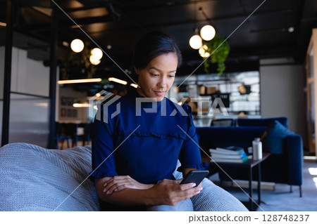 Asian woman wearing navy blouse sitting on light grey couch using smartphone in office lounge 128748237