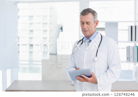 Mature male doctor wearing lab coat reviewing tablet in clinic with stethoscope at desk, copy space 128748246