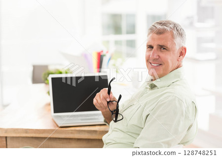 Senior man sitting at home office holding eyeglasses and smiling by laptop and folders, copy space Senior man sitting at home office holding eyeglasses and smiling by laptop and folders, copy space 128748251