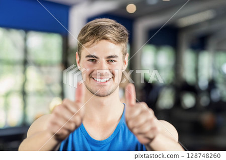 Man wearing blue tank top smiling, giving thumbs up while standing in gym with weight machines Man wearing blue tank top smiling, giving thumbs up while standing in gym with weight machines 128748260