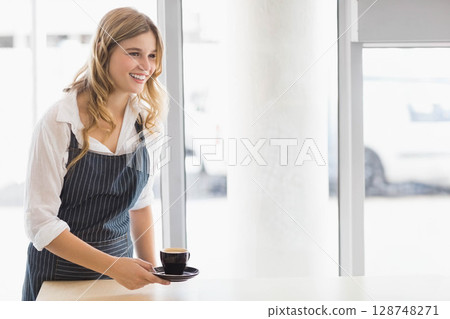 Female barista wearing apron serving coffee behind wood counter in cafe with black cup, copy space 128748271