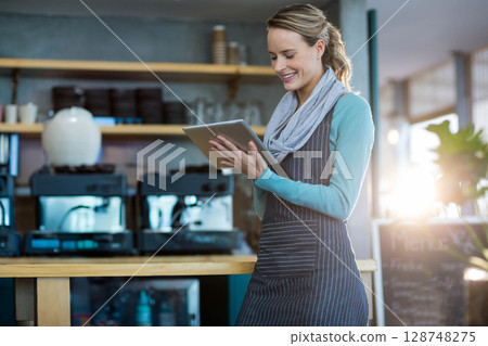 Female barista wearing apron tapping tablet behind counter in cafe with coffee machines, copy space 128748275