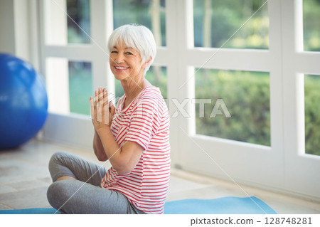 Senior woman wearing workout clothes sitting on blue mat beside exercise ball at home near windows Senior woman wearing workout clothes sitting on blue mat beside exercise ball at home near windows 128748281