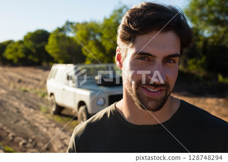 Man standing facing camera on dusty dirt track with white off-road vehicle behind, copy space Man standing facing camera on dusty dirt track with white off-road vehicle behind, copy space 128748294
