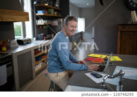 Mature adult man typing on laptop at central kitchen island with tablet calculator colorful papers Mature adult man typing on laptop at central kitchen island with tablet calculator colorful papers 128748295