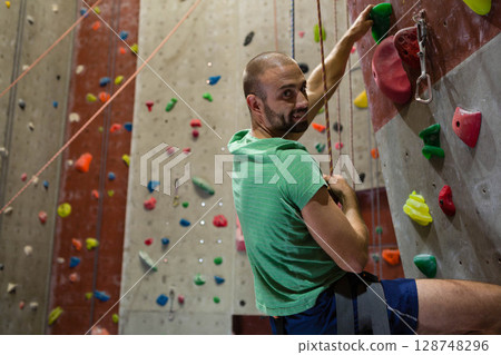 Man in his thirties climbing wall at climbing gym, wearing harness holding rope, copy space Man in his thirties climbing wall at climbing gym, wearing harness holding rope, copy space 128748296