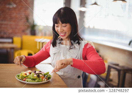 Asian woman sitting at wooden table in cafe, cutting salad on plate with fork and knife Asian woman sitting at wooden table in cafe, cutting salad on plate with fork and knife 128748297
