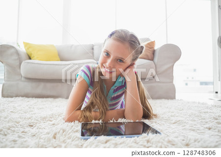 School-age girl lying on plush white rug in bright living room gazing at tablet 128748306