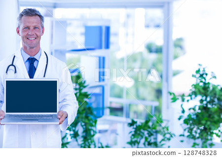Middle-aged male doctor holding open laptop toward viewer in clinic with stethoscope, copy space 128748328